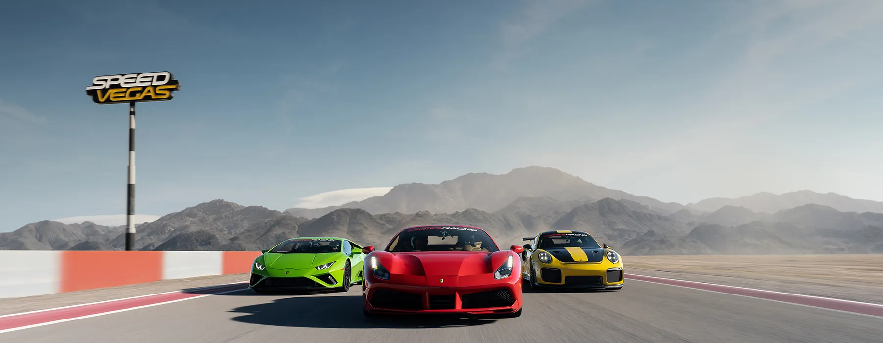 Three sports cars speed on a racetrack against a mountain backdrop. A sign reads